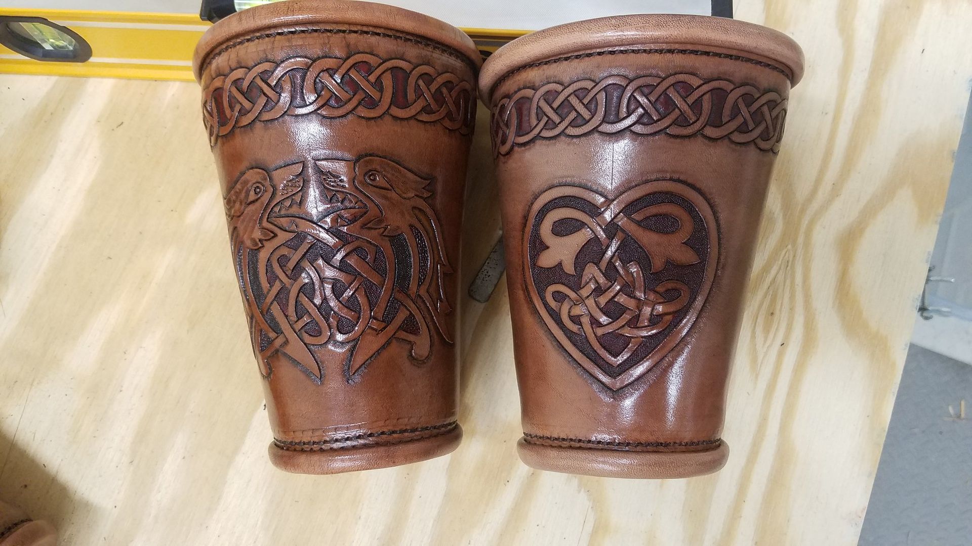 Two carved brown leather cups with Celtic-style patterns on a light wooden surface