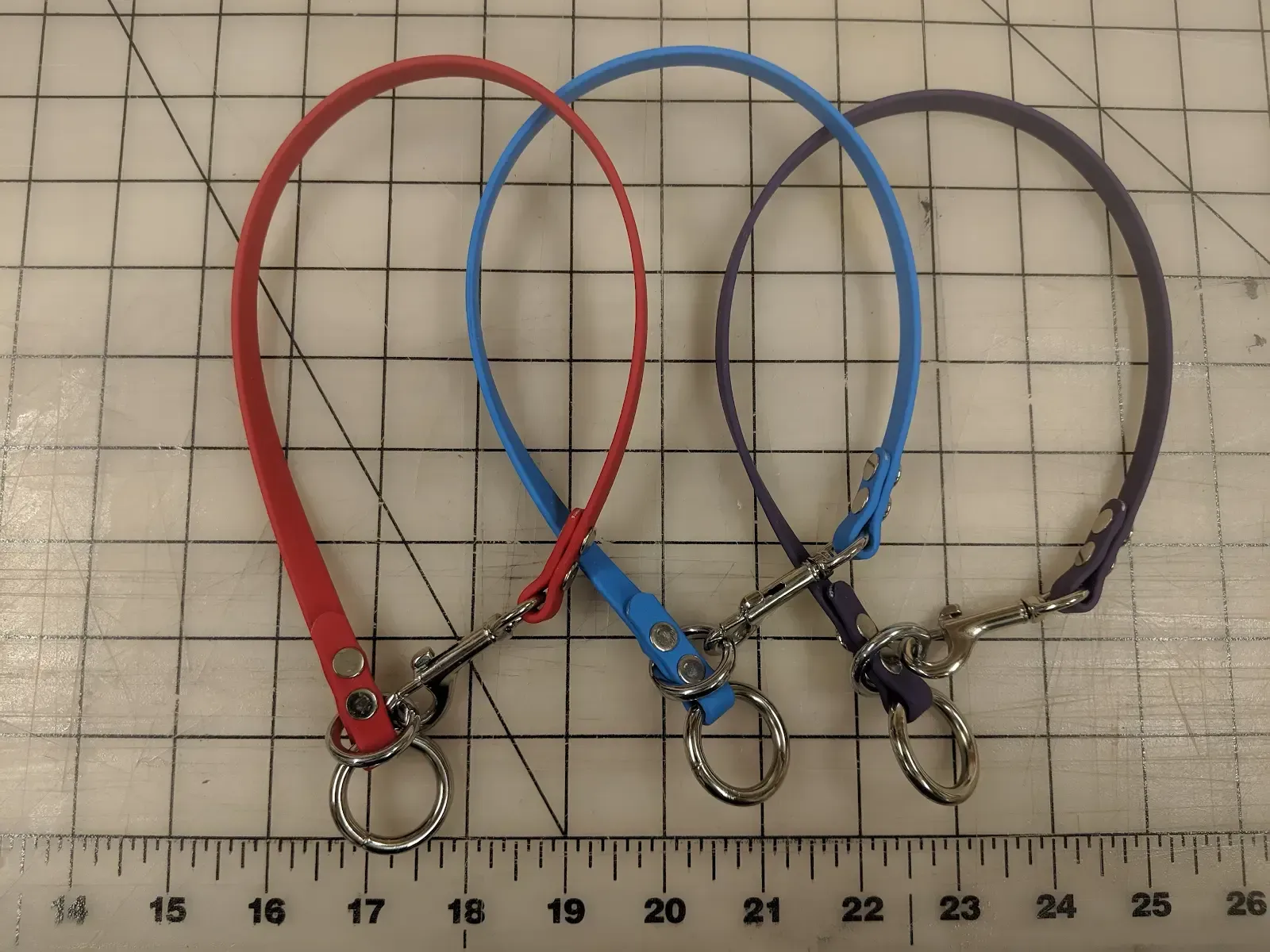 Three dog collars with metal clips, in red, blue, and black, laid side by side on grid paper.