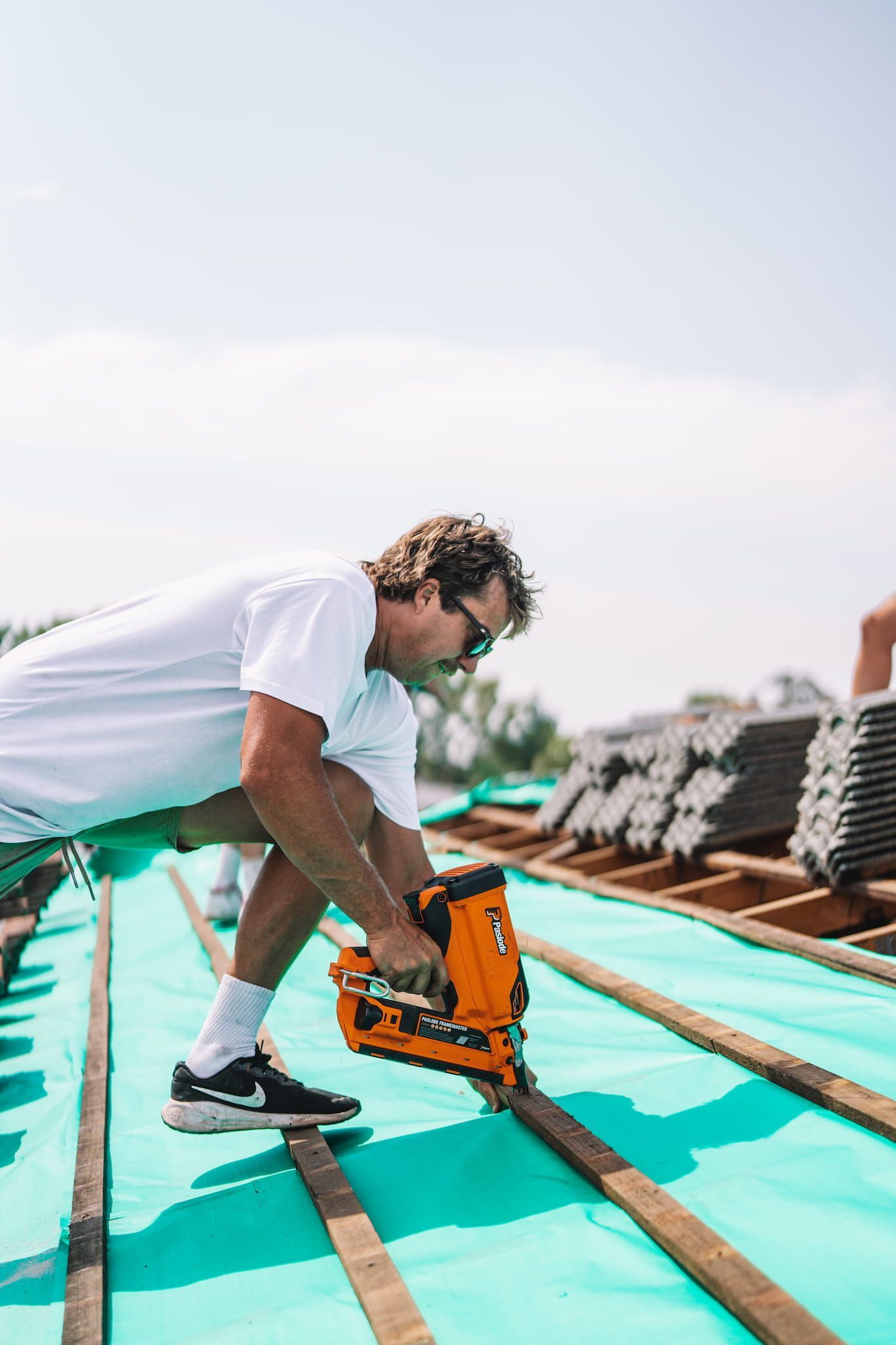 Roofer using a nail gun on a roof, sunny day. He's wearing a white shirt and sunglasses.