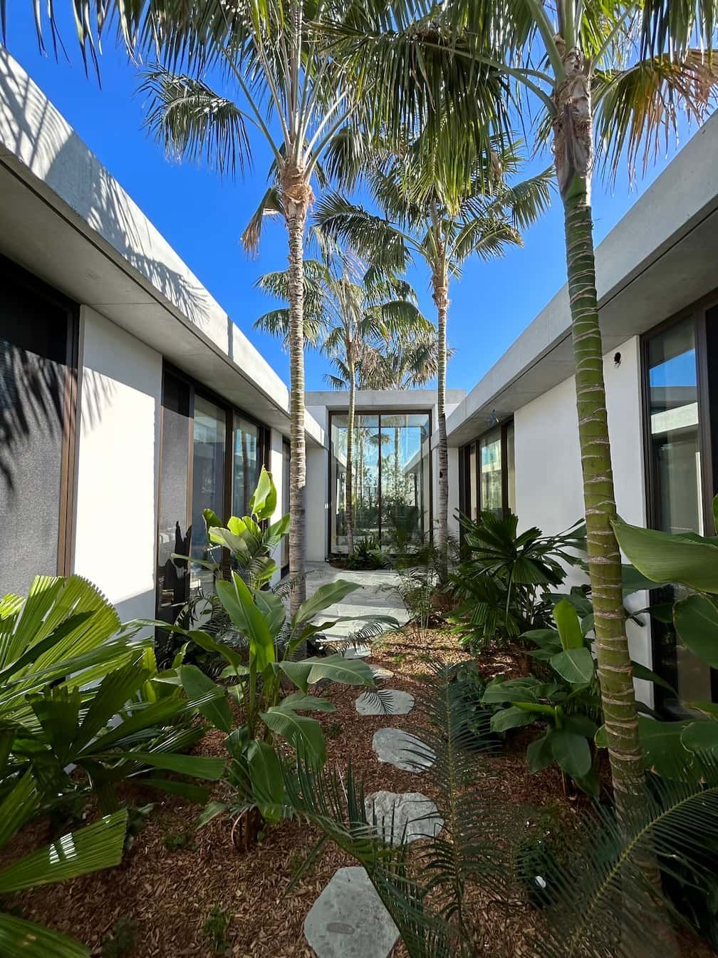 Courtyard with stone path, lush plants, and palm trees between two buildings under blue sky.