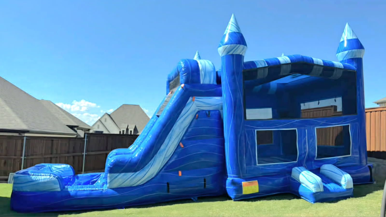 Big Blue A blue and white inflatable bounce house with an attached slide, situated on a grassy lawn in a suburban backyard.