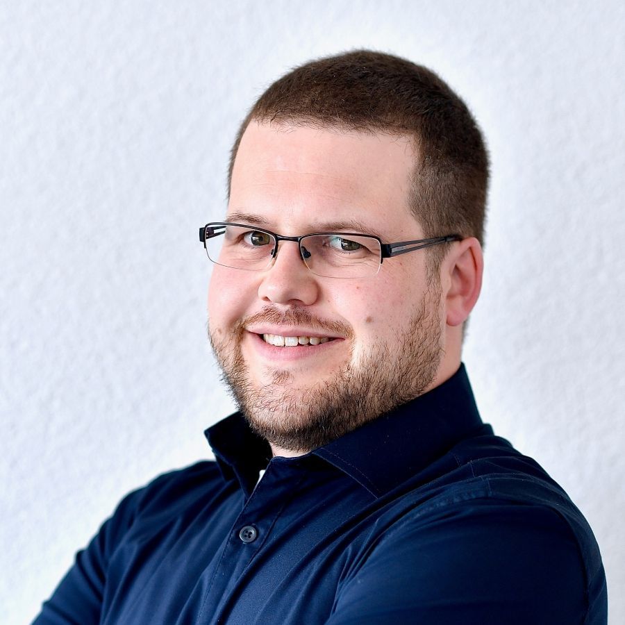 Ein Mann mit blauem Hemd steht vor einer weissen Wand
