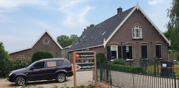 Een bruine bakstenen boerderij met een donker dak en een kleine auto ervoor geparkeerd, met een bord in een grindoprit.