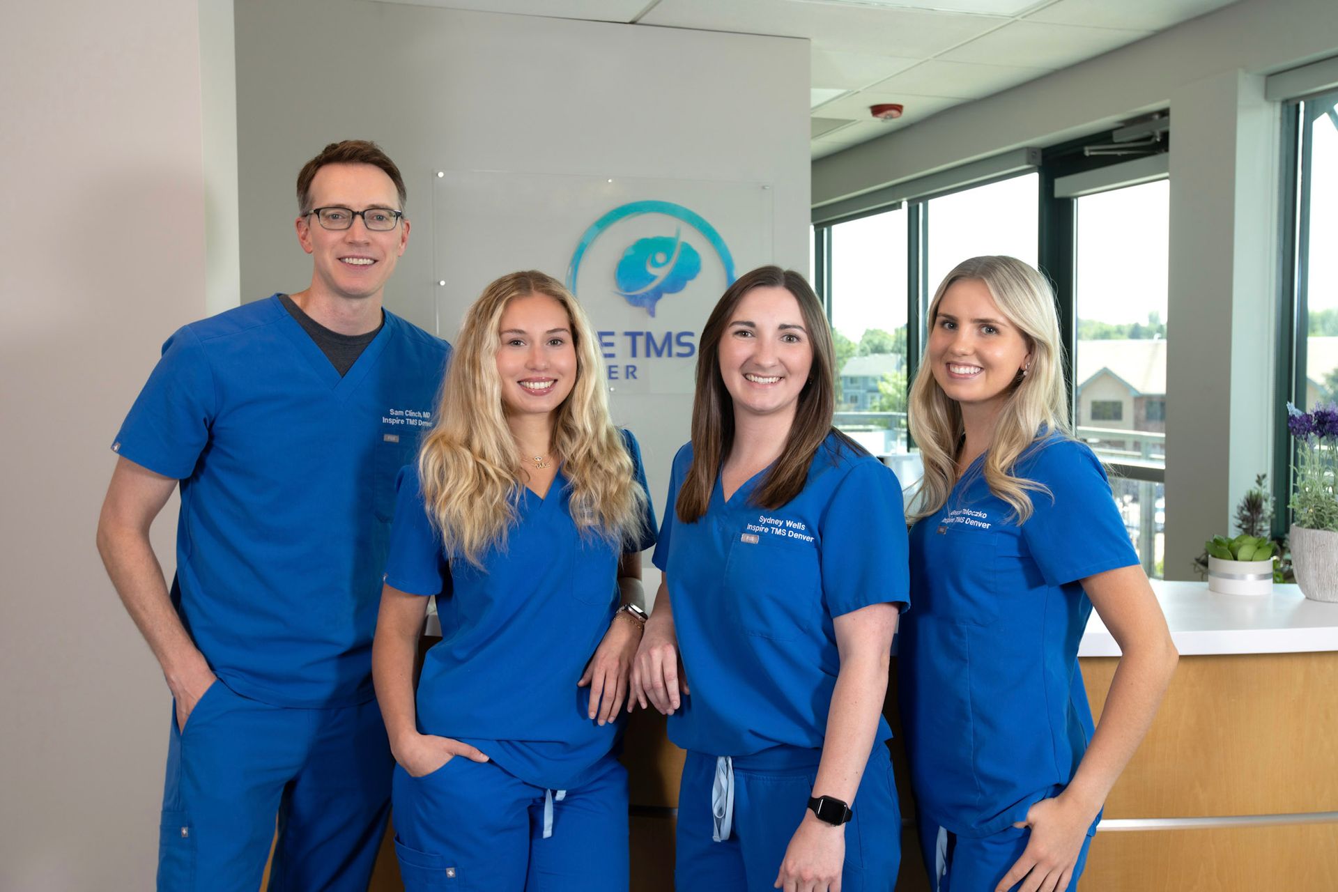 Four medical staff members in blue scrubs smiling while standing in front of a clinic logo.