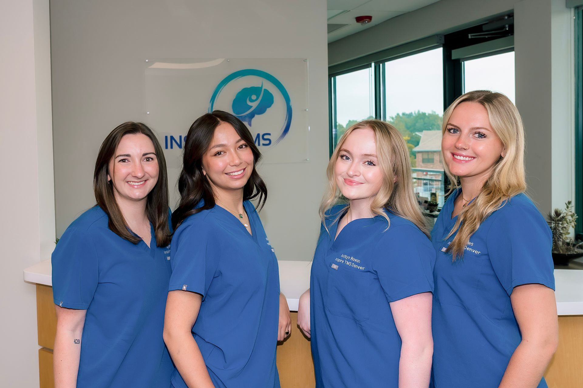 Four healthcare staff in blue scrubs standing together in a clinic hallway, smiling at the camera.