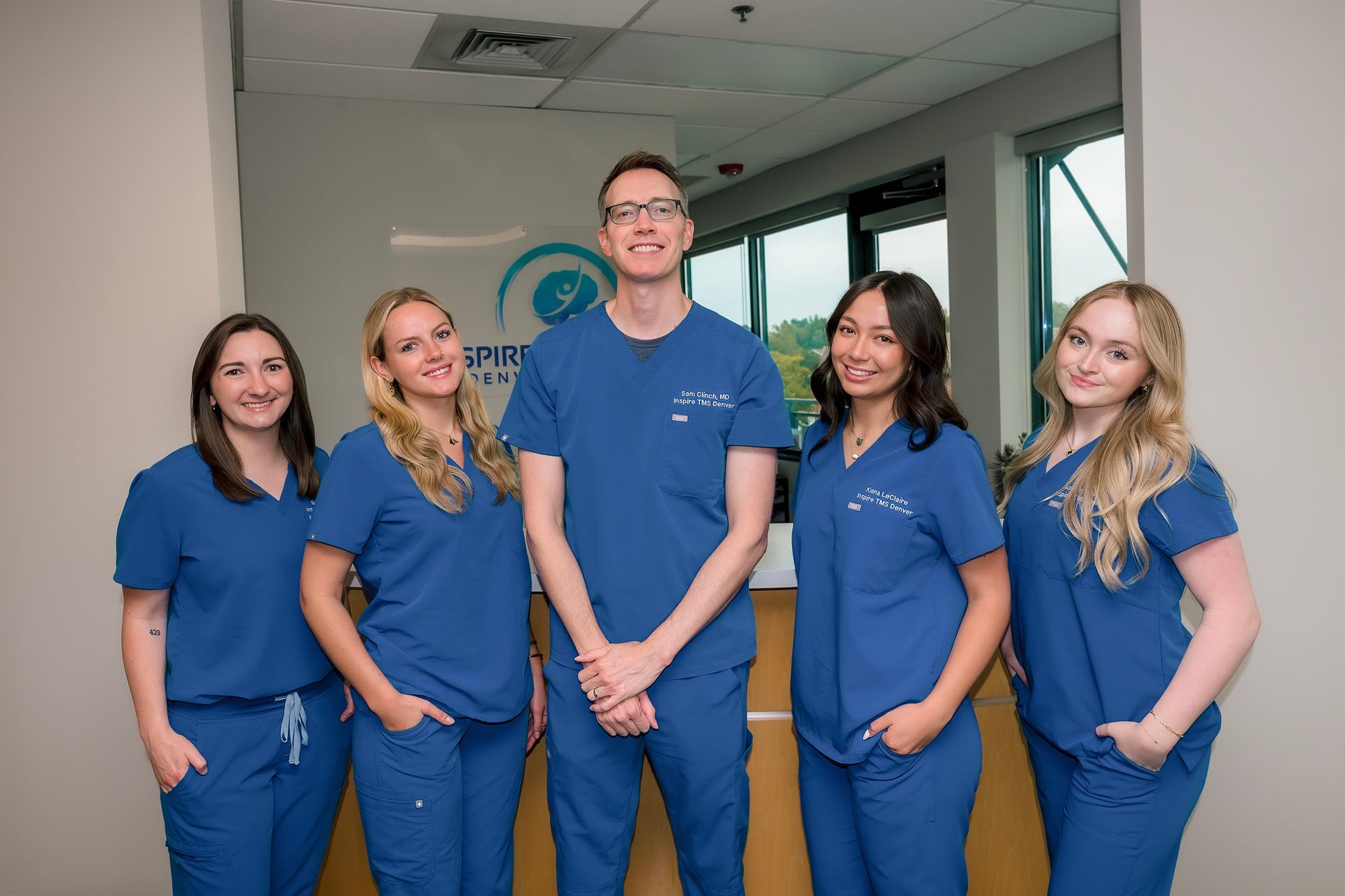 Five medical staff in blue scrubs standing in a clinic hallway, smiling at the camera.