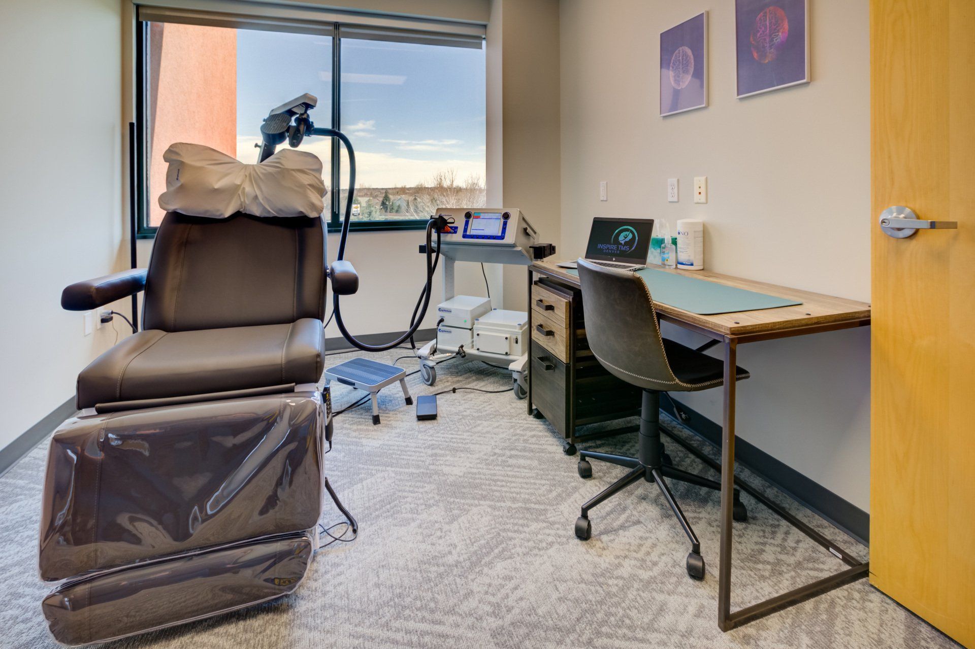 Medical exam room with a reclining treatment chair, desk, computer, and medical equipment by the window