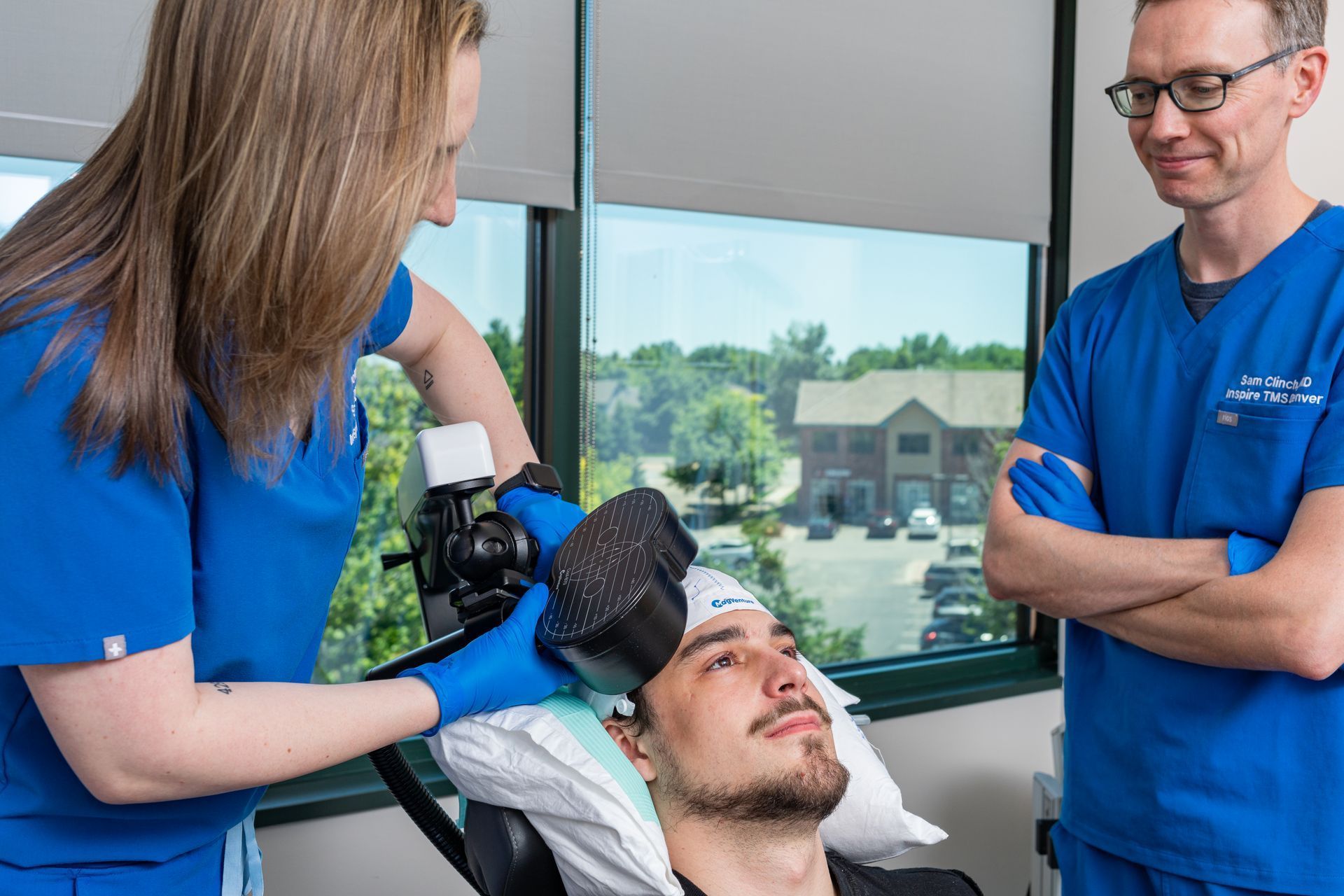 Two medical professionals in blue scrubs administer a TMS therapy treatment to a patient sitting in a reclining chair.