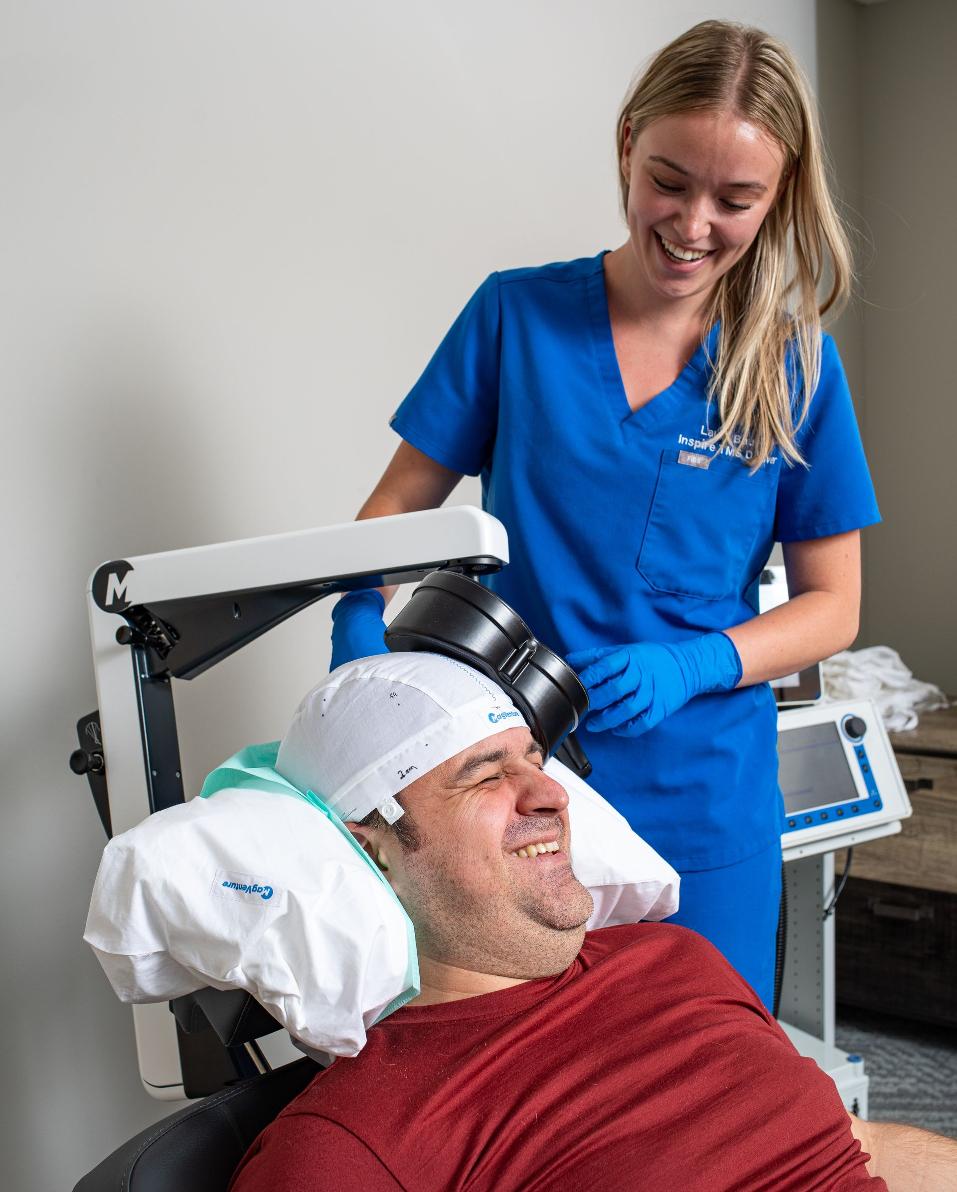 Clinician in blue scrubs adjusts a scalp treatment device on a smiling patient in a clinic chair