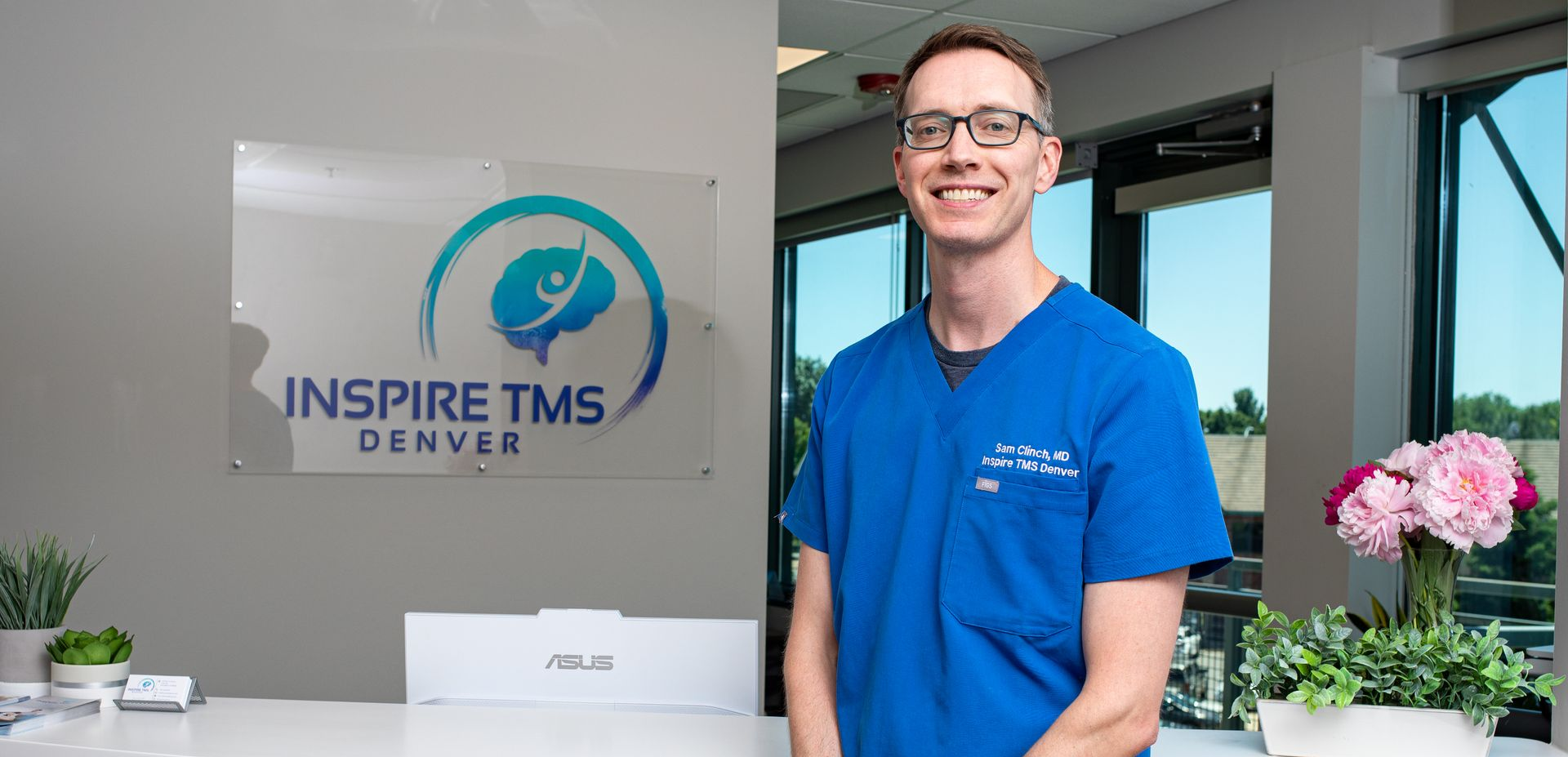 A smiling medical professional in blue scrubs stands in front of an Inspire TMS Denver sign in an office setting.