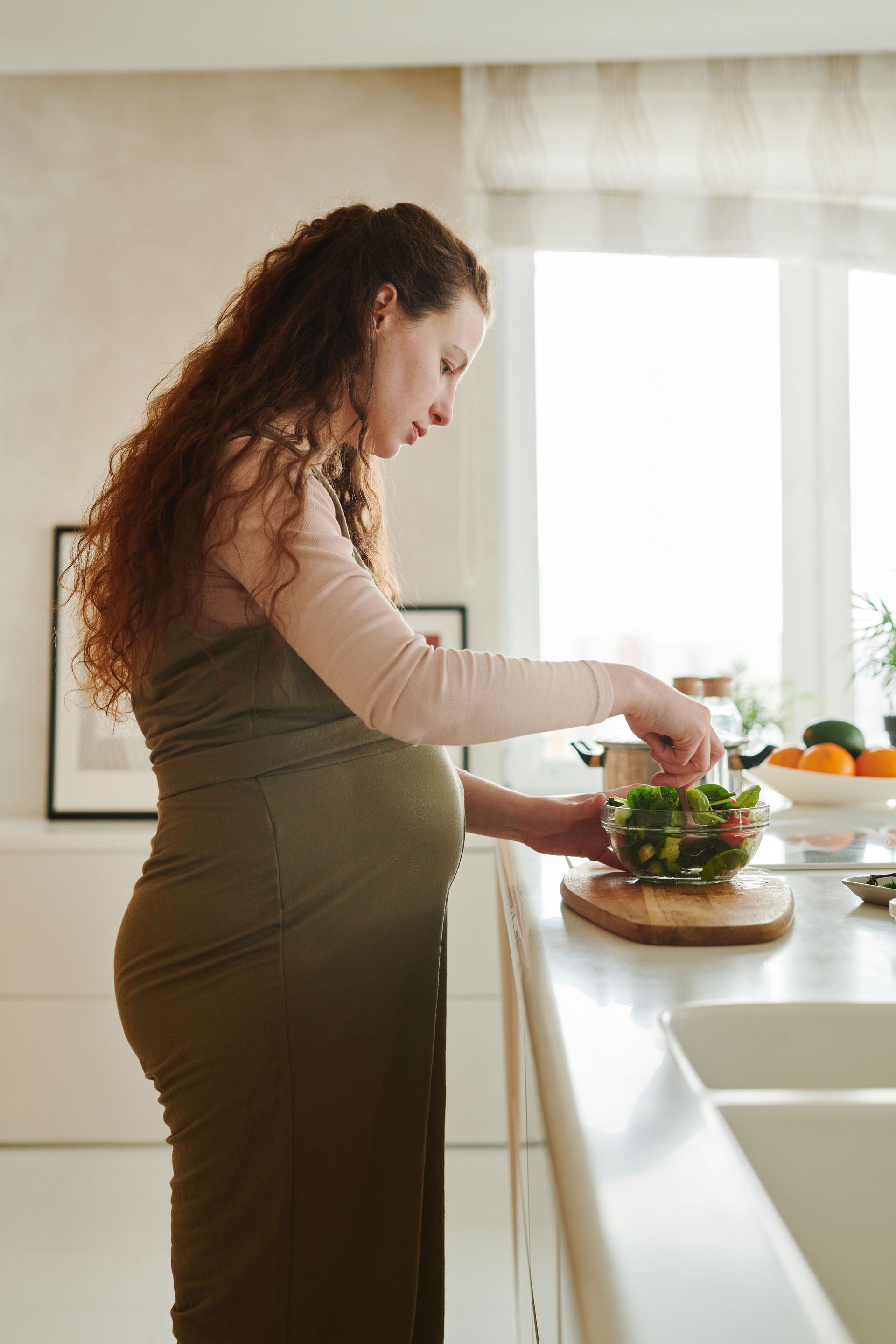Pregnant woman preparing a salad in a bright kitchen by the window