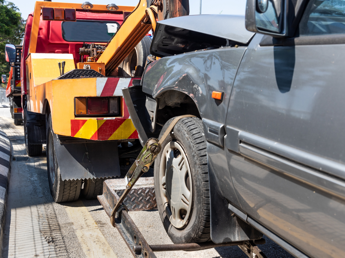 Tow truck towing a damaged gray car on a sunny day.