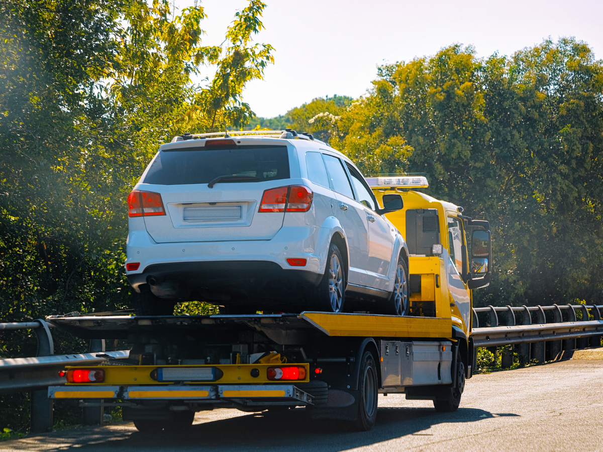 White SUV loaded onto a yellow tow truck on a road with green trees in the background.