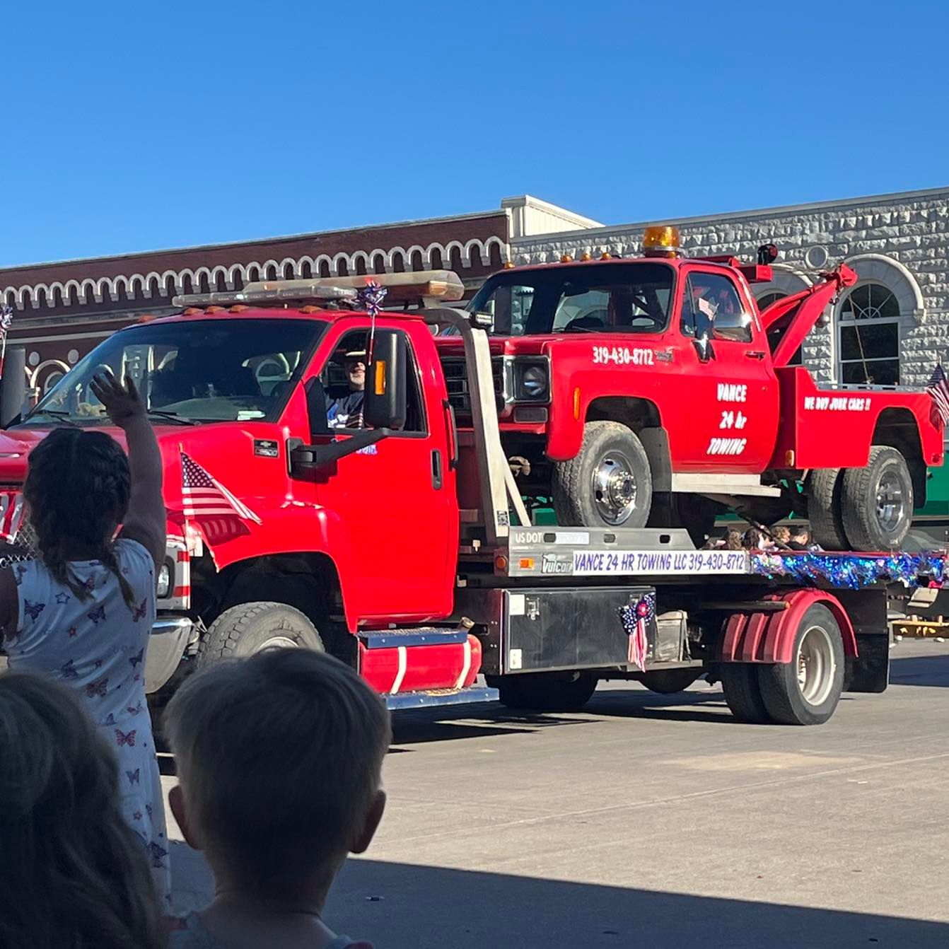 Red tow truck in parade carrying another red truck, waving children.
