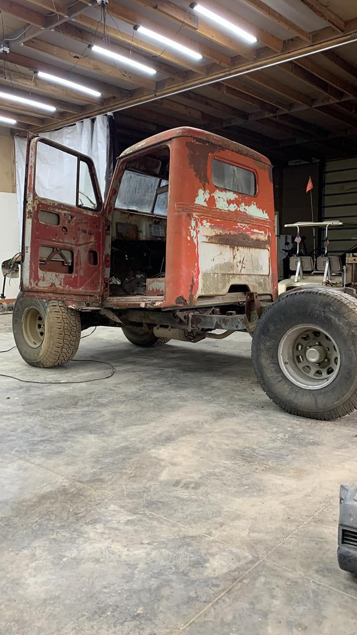 Rusting red truck cab on chassis, missing wheels and components, in a garage.
