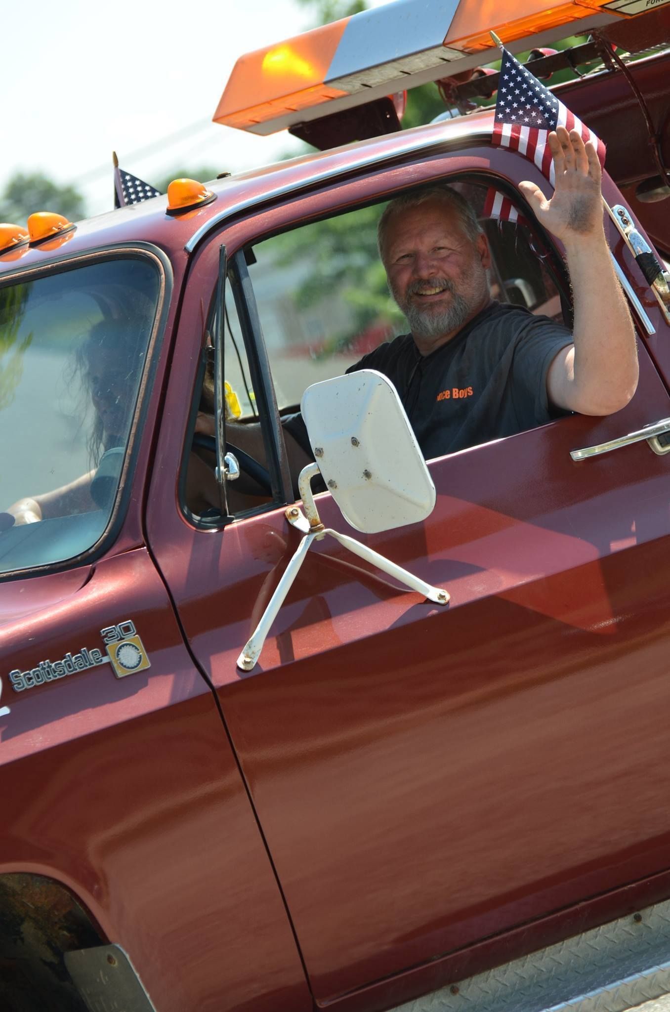 Man waving from a maroon tow truck, holding an American flag; orange lights on the roof.