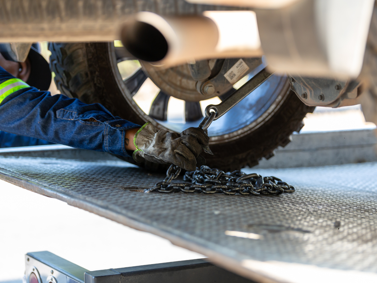 Person securing a vehicle with chains on a tow truck platform, near tire and exhaust.