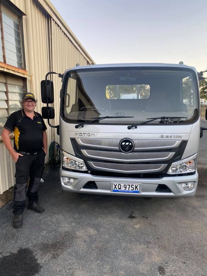 Worker stands beside a silver Foton truck in an industrial yard.