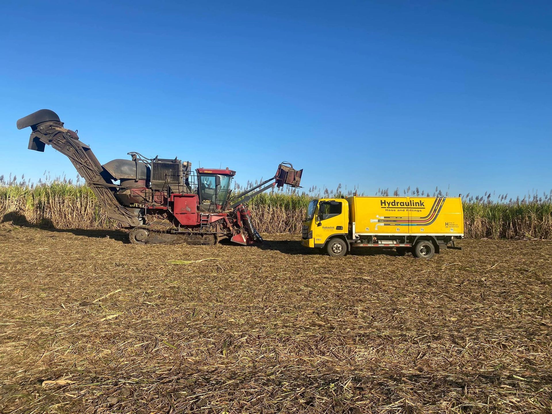 Yellow truck beside a corn harvester in a harvested field under a clear blue sky