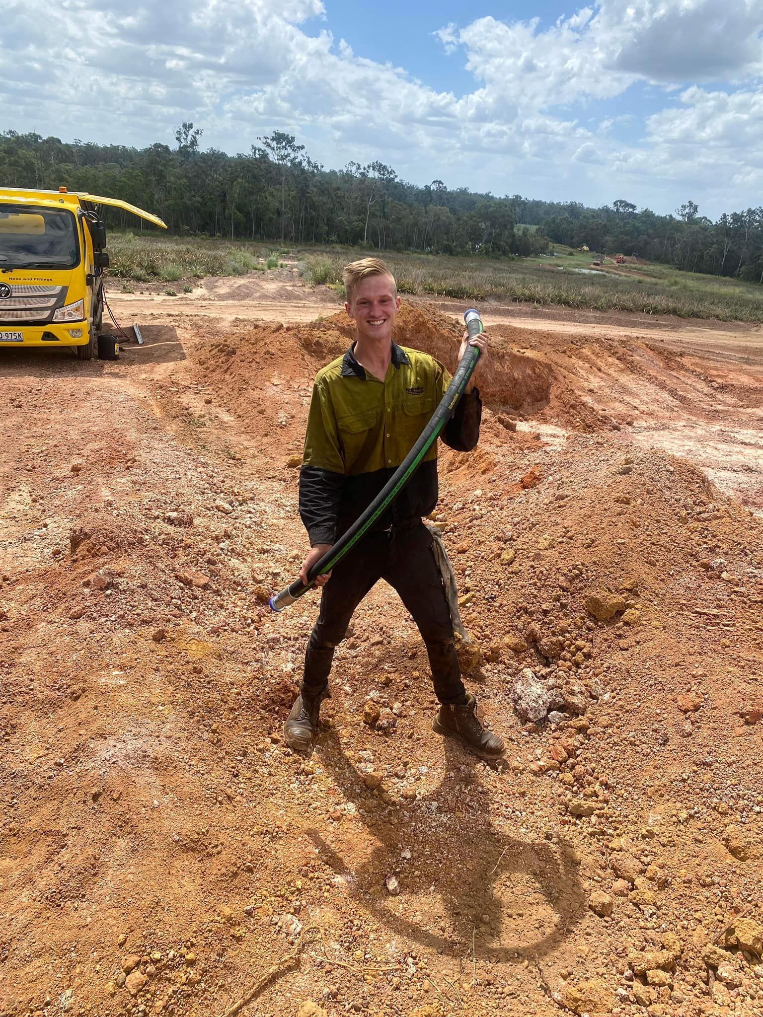 Man holding a long tool on a muddy construction site, with a yellow excavator in the background.