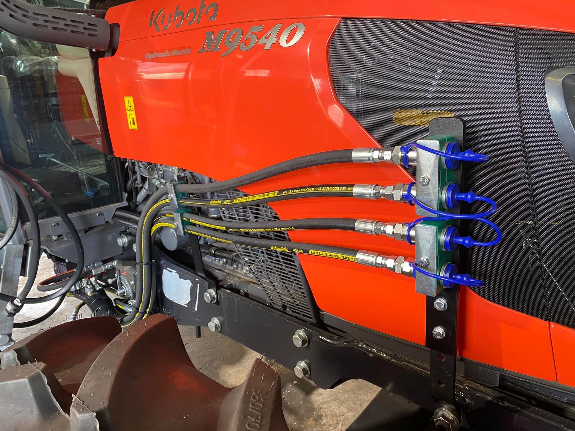 Close-up of a red agricultural machine with four blue hydraulic hoses and a black patterned side panel