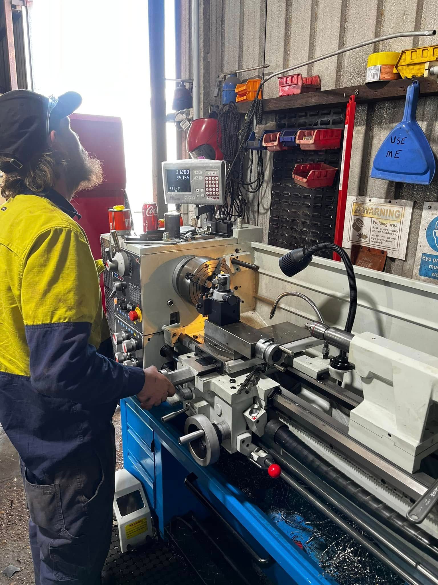 Worker in a yellow shirt operating a lathe in a machine shop, with tools and shelves in the background.