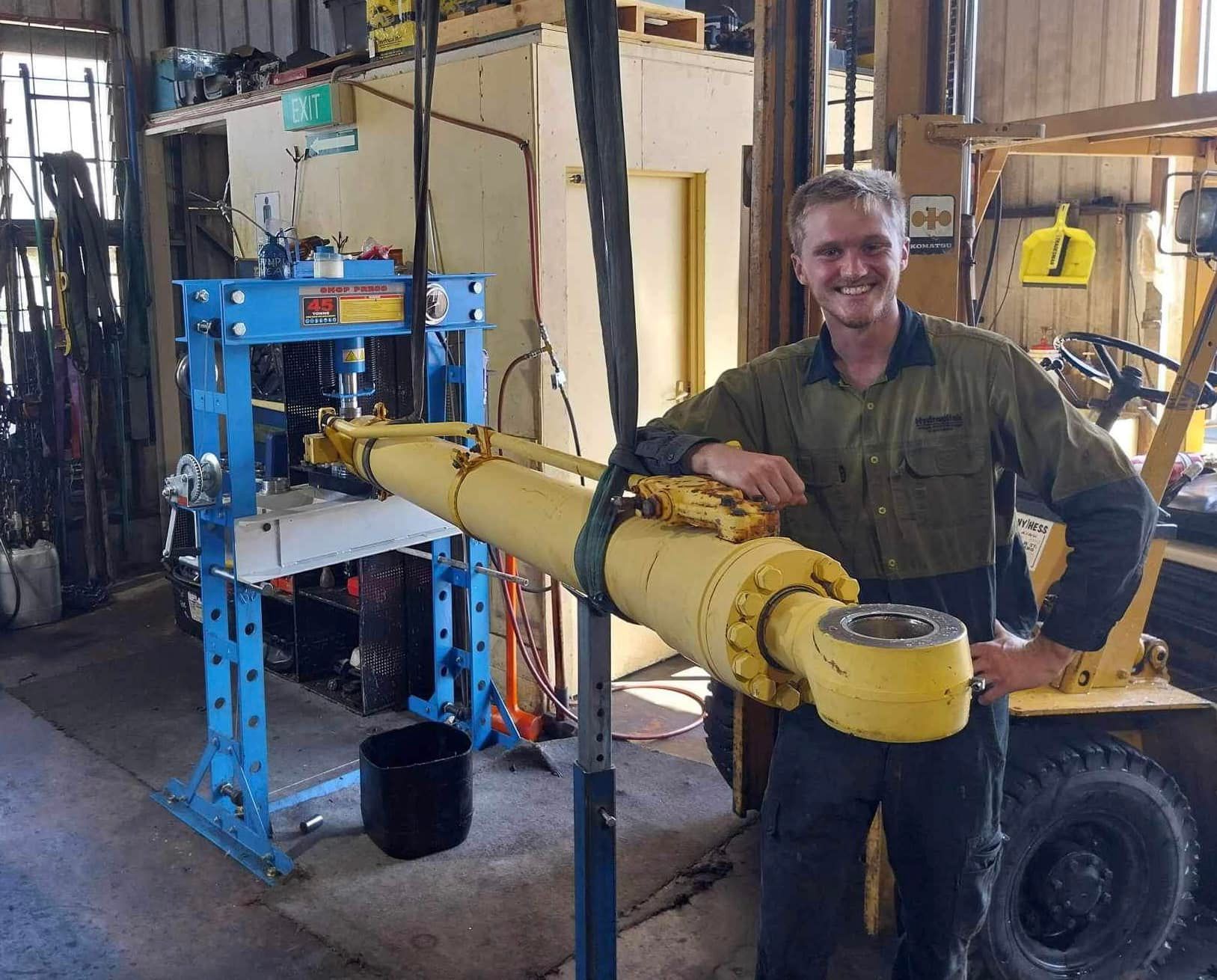 Man smiling beside a large yellow machine arm in a workshop, with blue metal equipment in the background