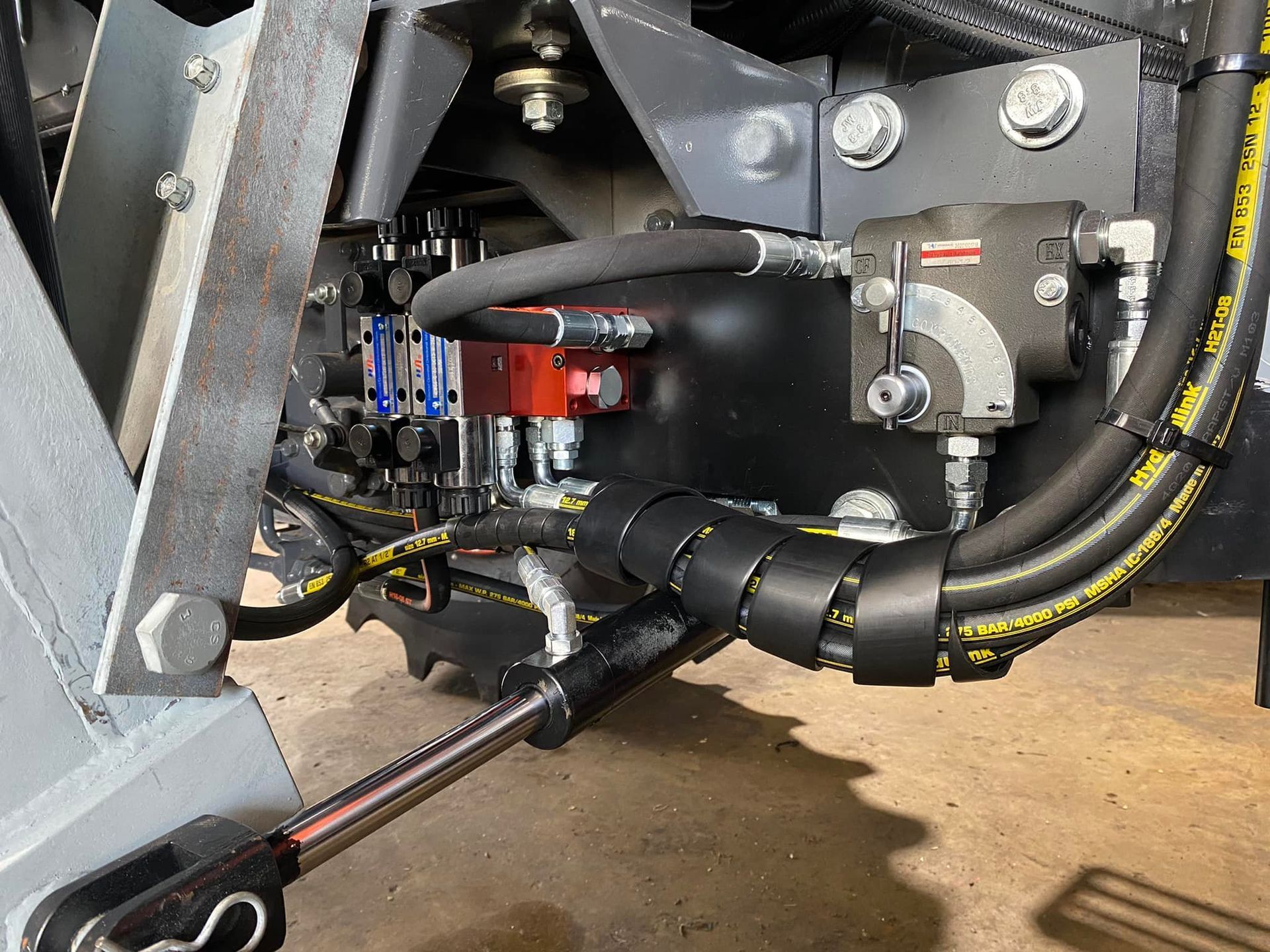 Close-up of machine undercarriage with hoses, wiring, and a red component on a metal frame.