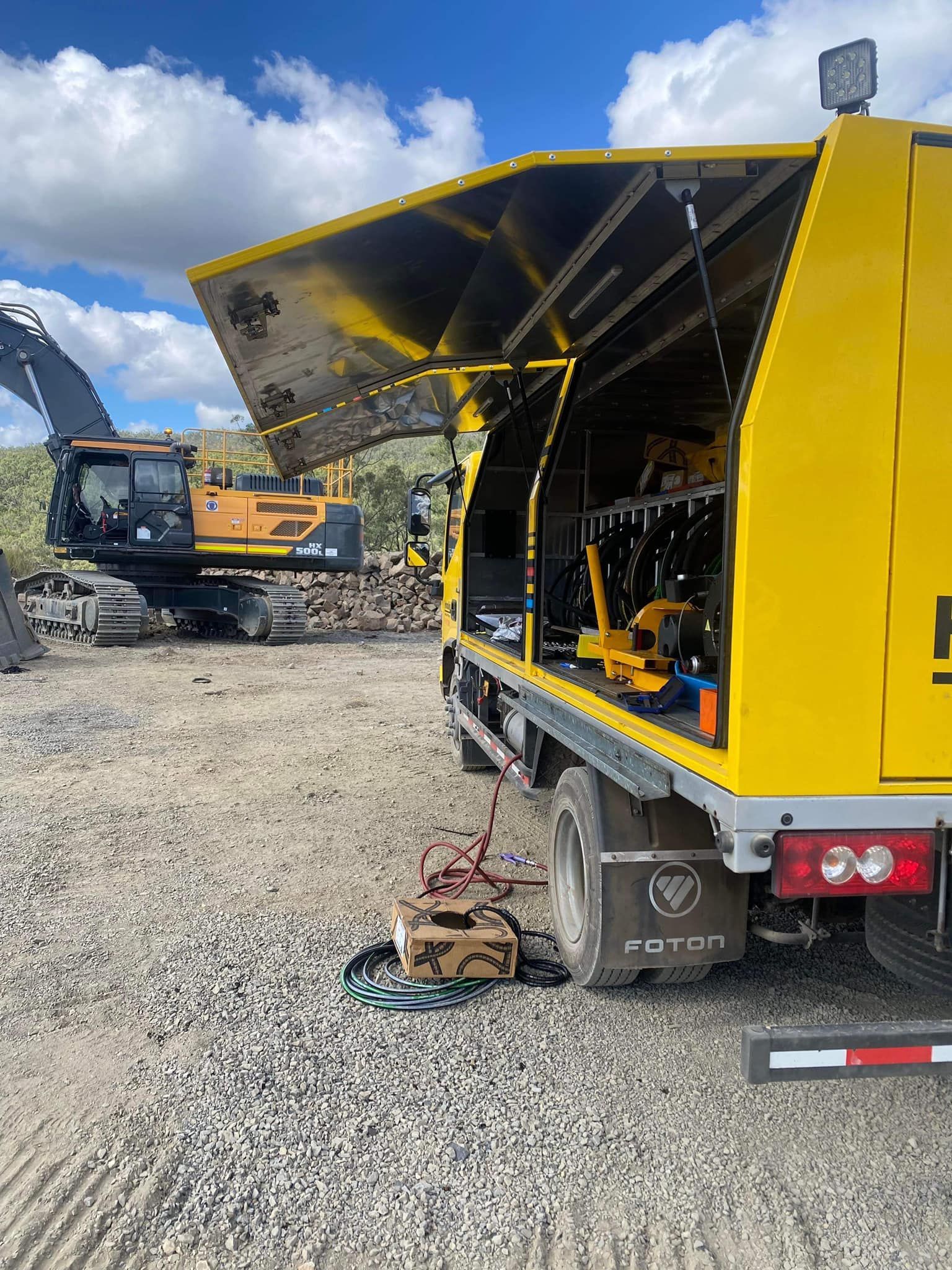 Yellow service truck with open side compartment at a gravel construction site, excavator in background