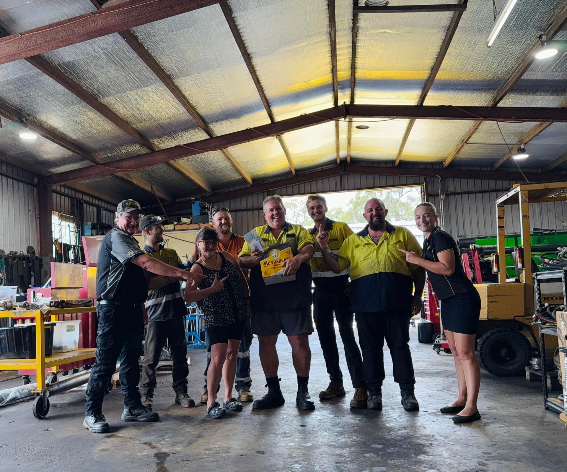 Six workers in yellow safety shirts stand in a warehouse, holding a trophy and posing for a group photo.