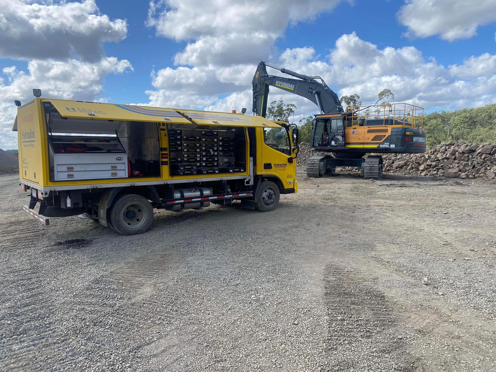 Yellow utility truck beside an excavator on a gravel worksite under a cloudy sky