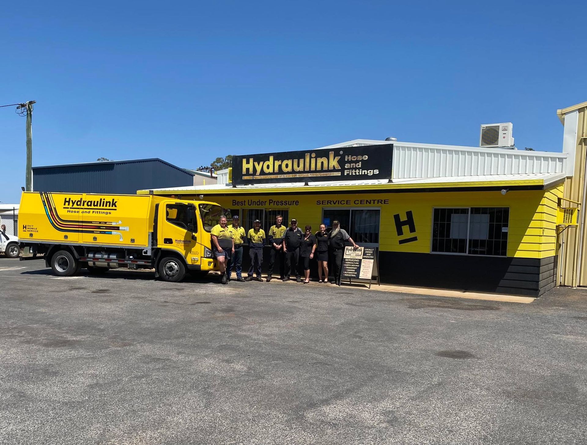 Yellow HydraLink truck and group of people standing outside a yellow-and-black industrial building on a sunny day