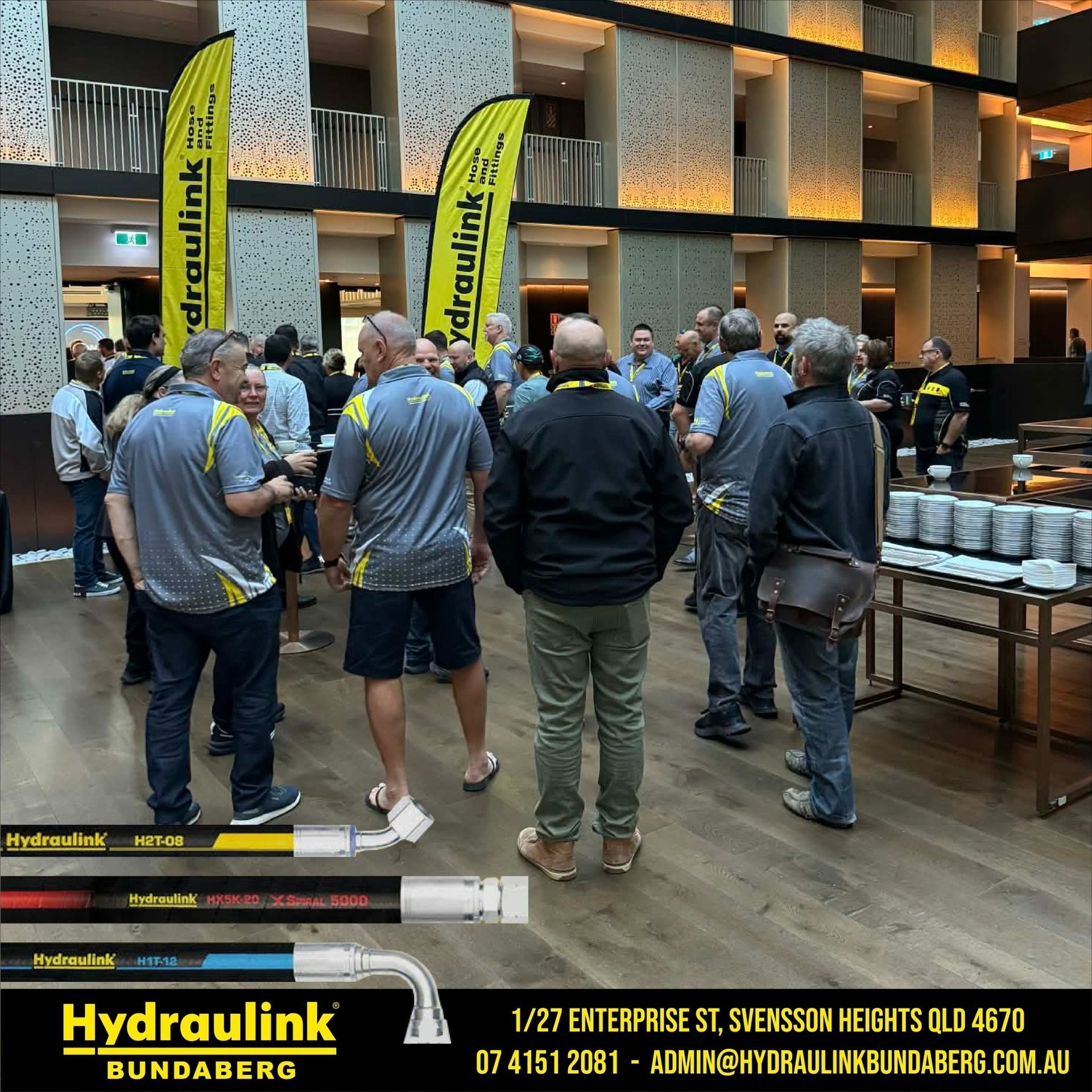 People gathered in a building lobby at a Hydraulink event beside display tables and banners.