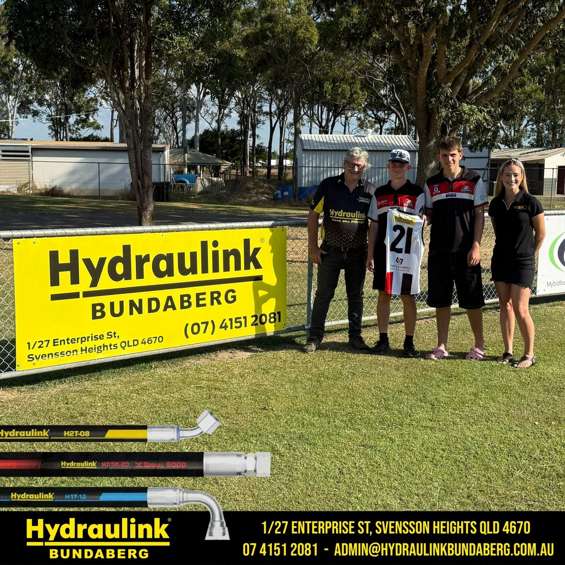 Four people standing beside a Hydraulink Bundaberg sign on a grassy field, holding a number 21 jersey
