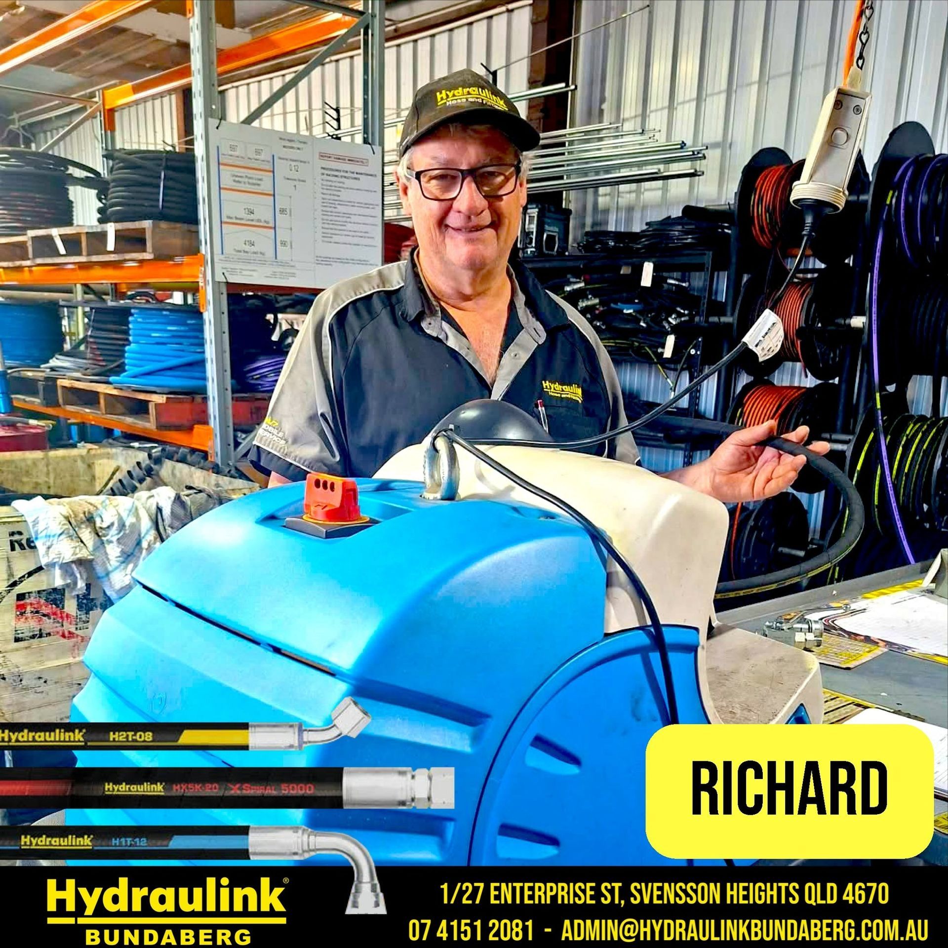 Man in a warehouse smiling beside a blue Hydraulink hose machine, holding a hose reel.