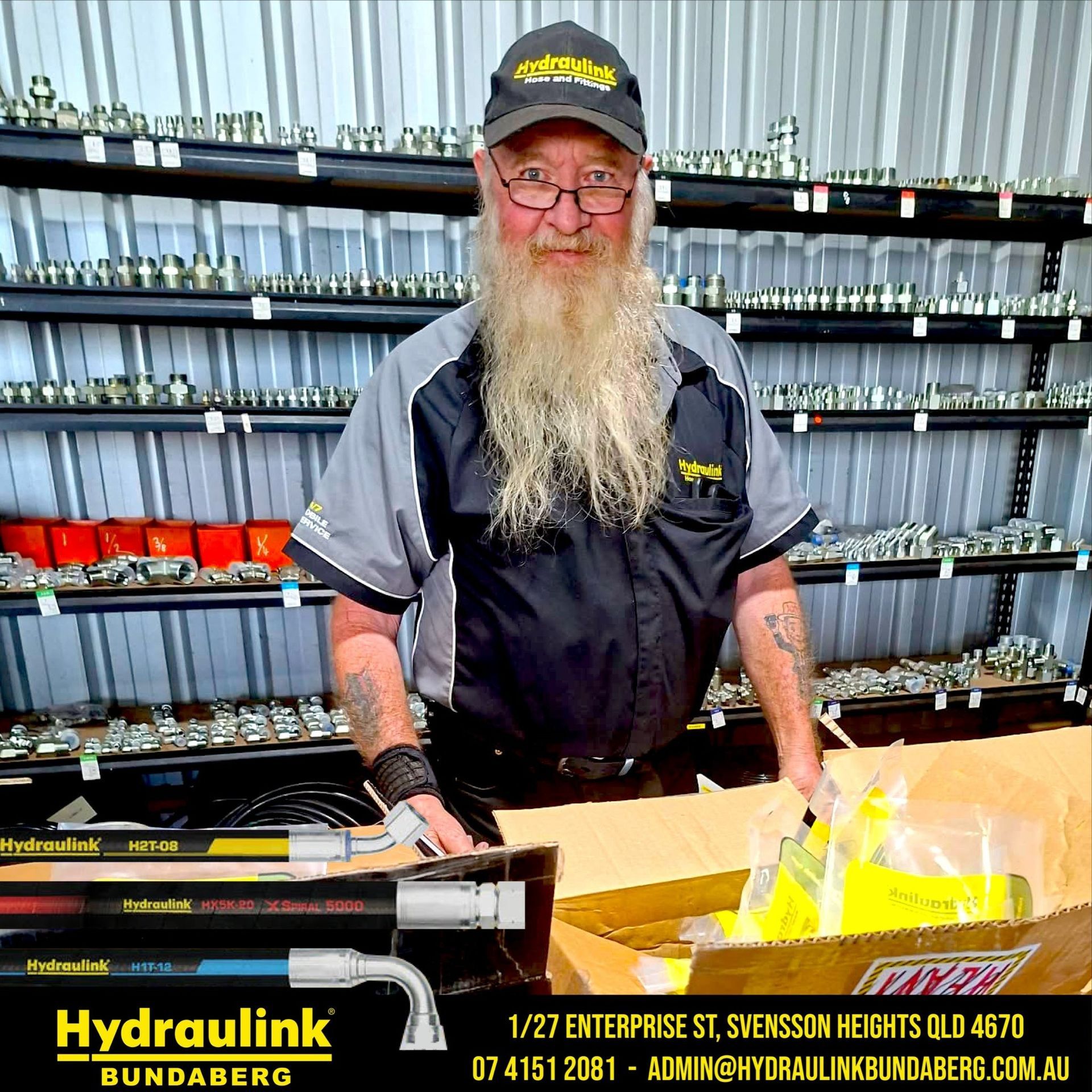 Man with long beard behind Hydraulink counter, shelves of fittings behind him, holding yellow plastic bags.