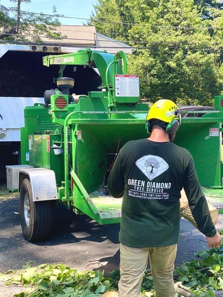 Worker in hard hat standing by a green wood chipper truck outdoors.