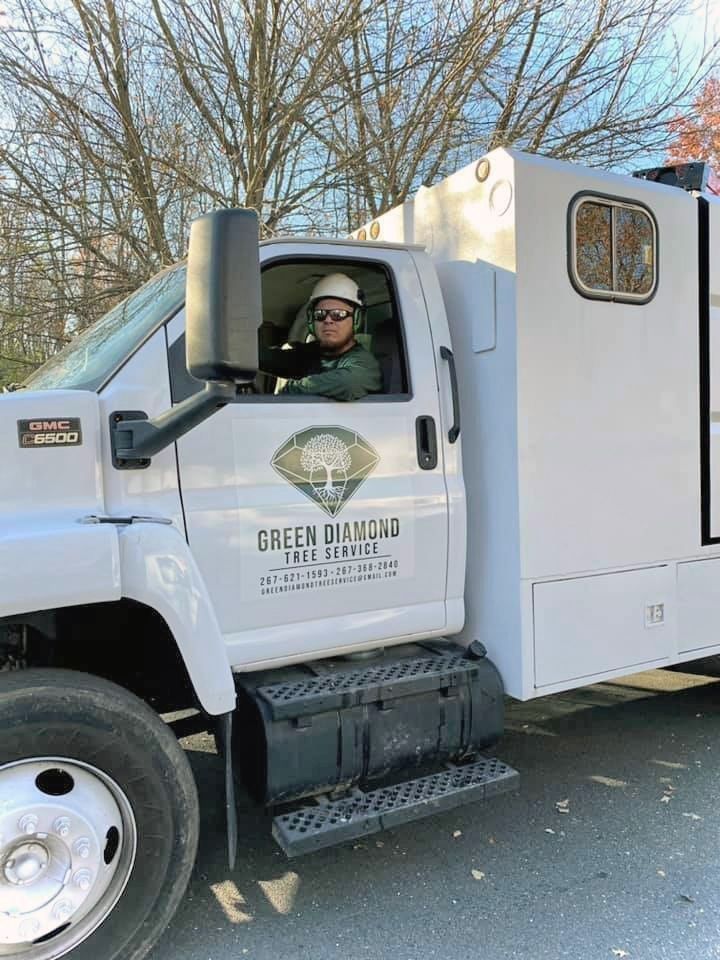 Driver in white utility truck beside trees, wearing sunglasses and a hat.