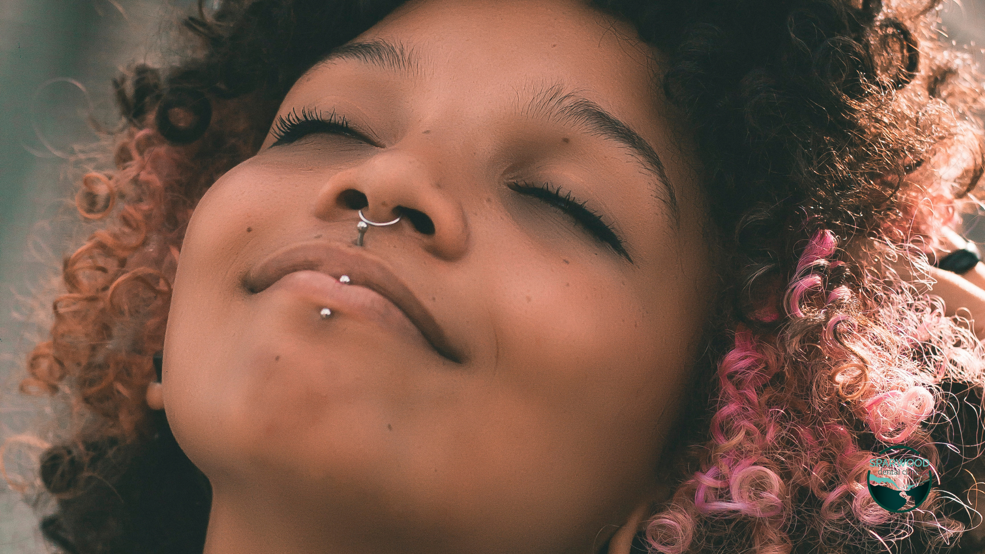 A woman with pink hair and a nose ring is smiling with her eyes closed.