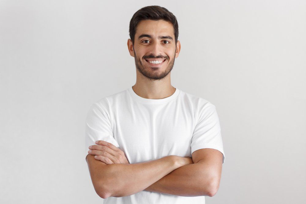 A man in a white t-shirt is standing with his arms crossed and smiling.