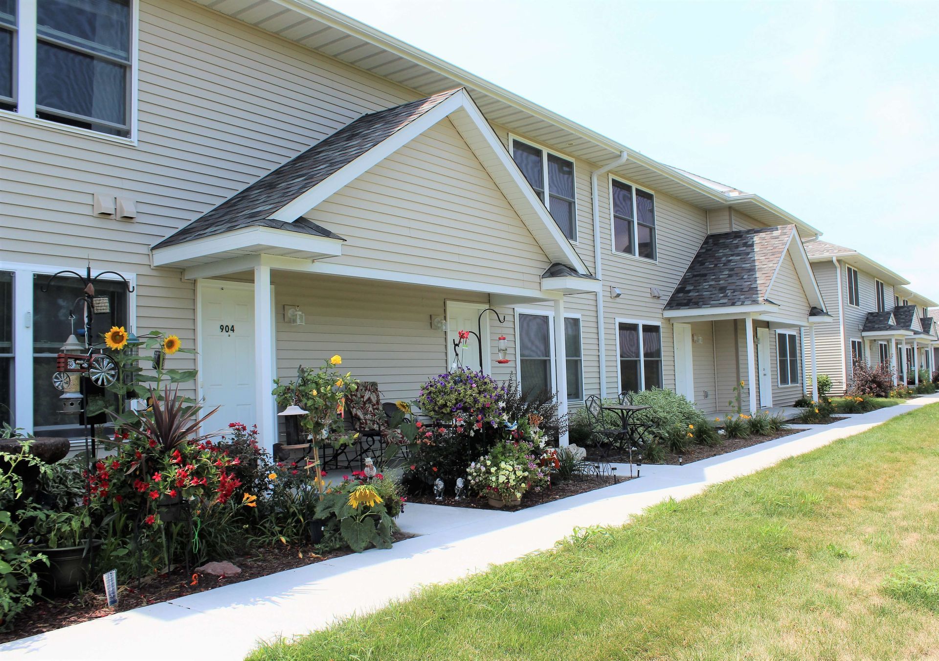 Townhomes with tan siding and individual entryways, a sunny day, a sidewalk with grass on the right, and flowerbeds.