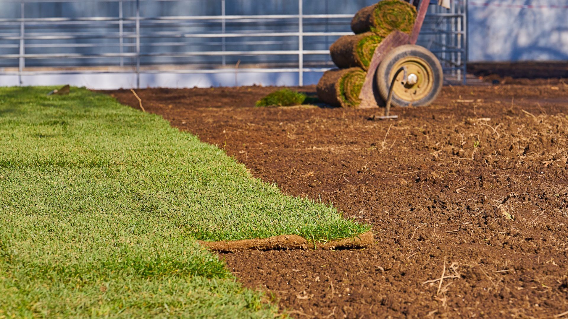 Freshly tilled brown soil beside a strip of green grass with a wheelbarrow near a fence