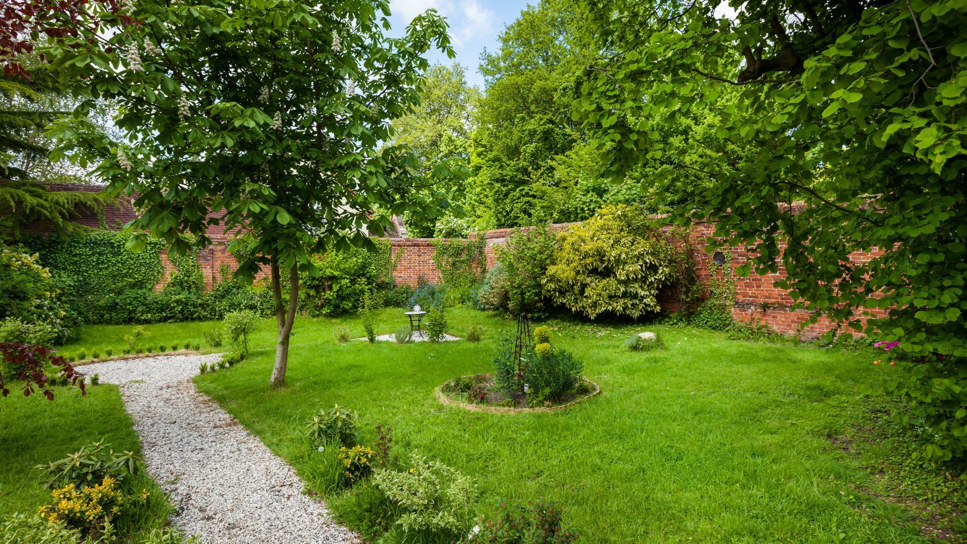 Lush green garden with a curved gravel path, trees, shrubs, and a small flower bed under a cloudy sky
