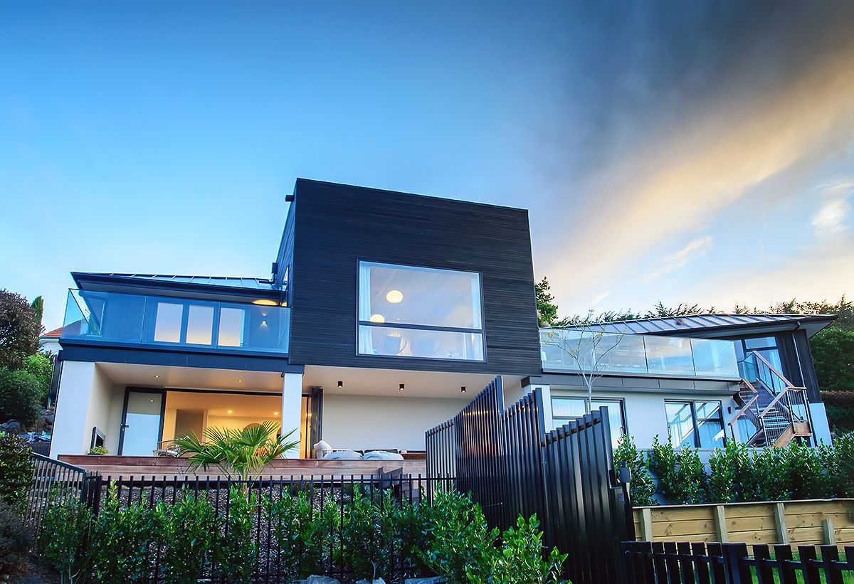 Modern two-story house with dark siding, lit windows, and a garden fence at dusk