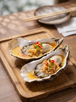 Two grilled oysters on a wooden board, topped with sauce and red and green garnish.