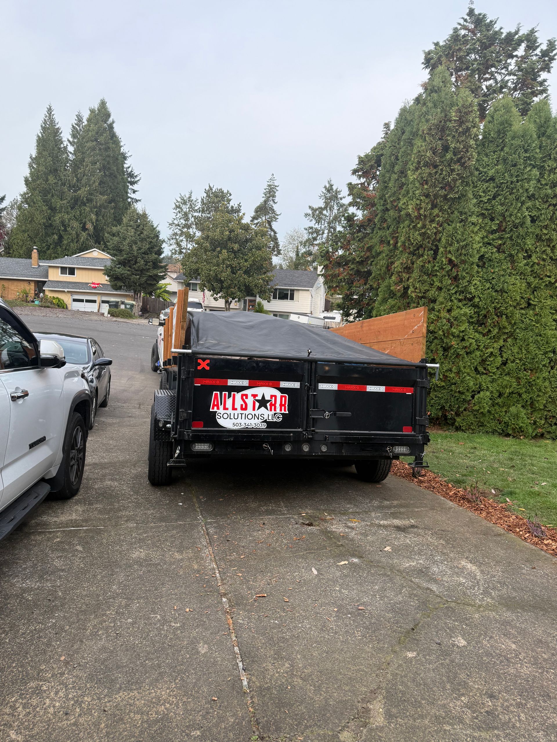 Black pickup truck parked in a gravel driveway beside a white vehicle, with trees and a hedge nearby.