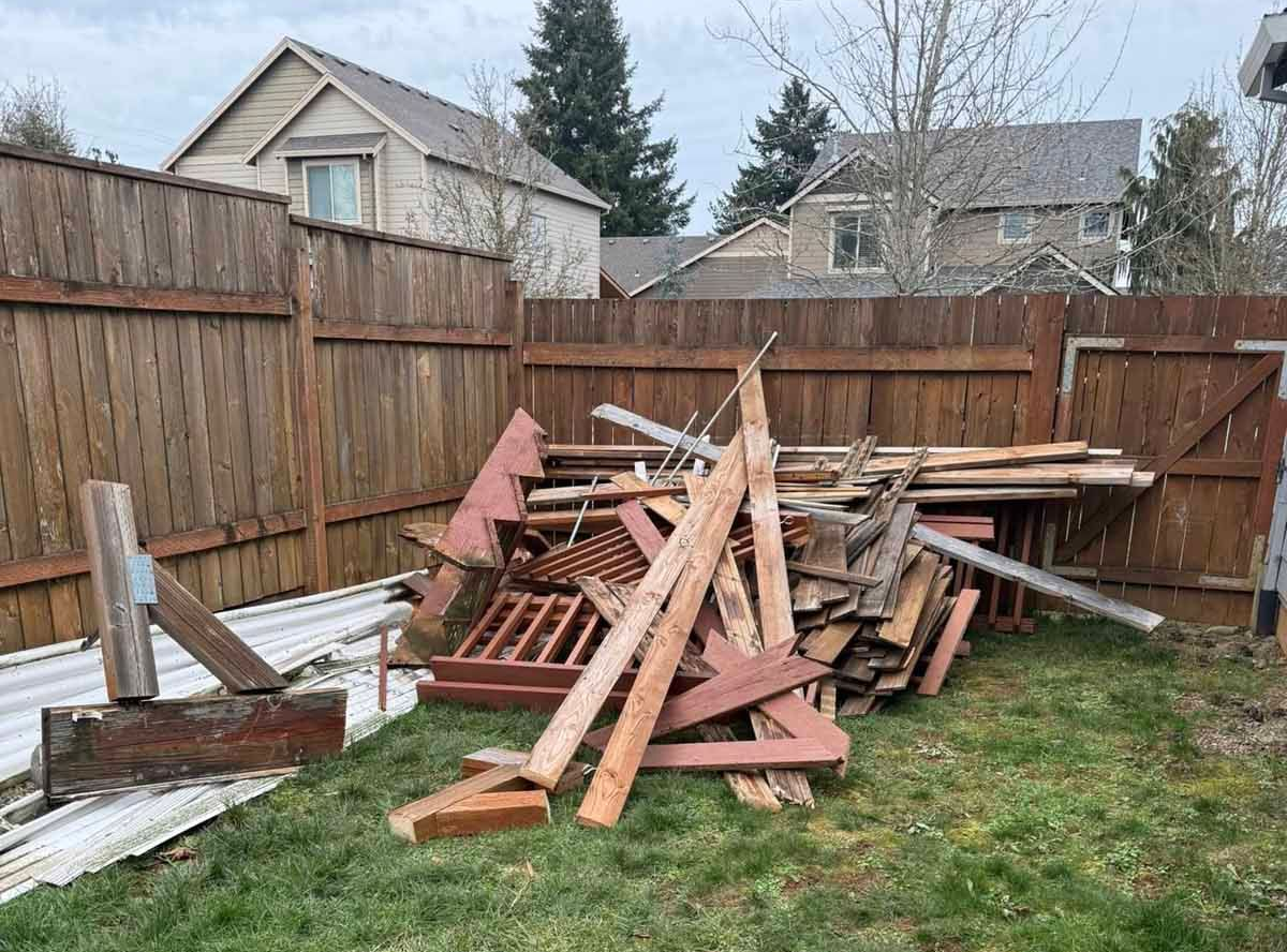 A large pile of discarded reddish-brown wooden boards and debris sits in a backyard corner against a tall wooden fence.