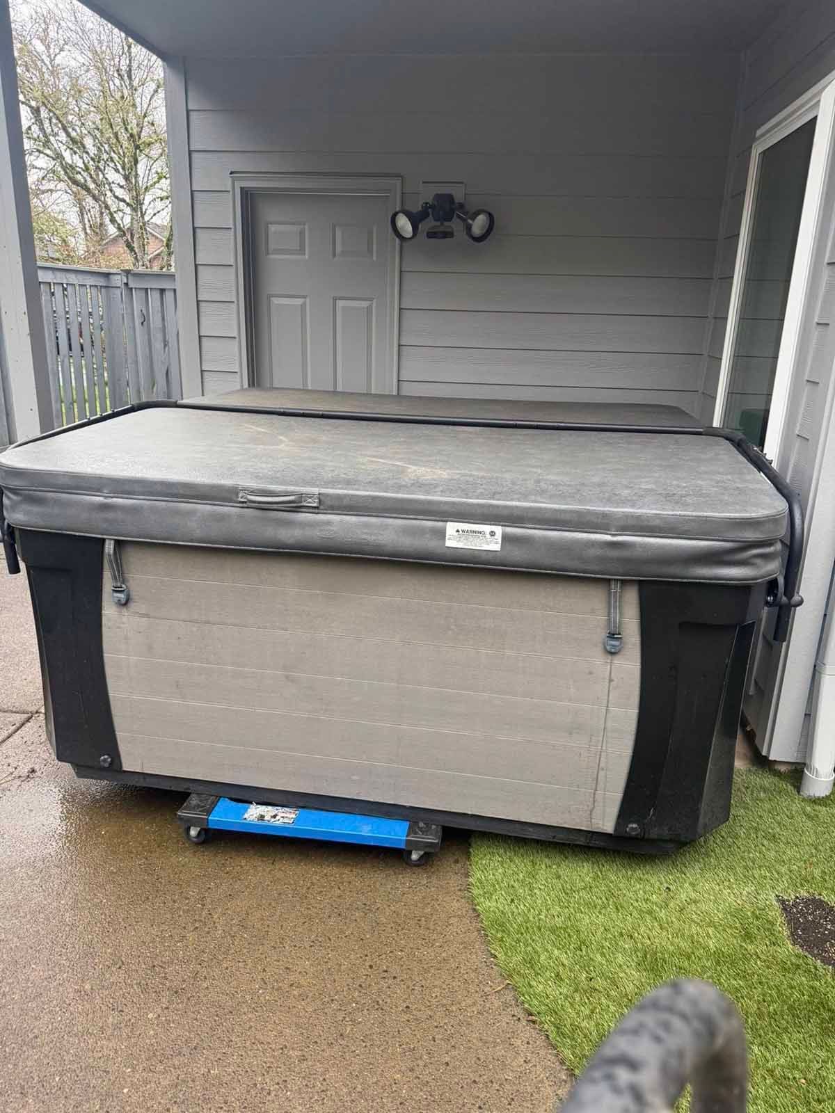 A covered hot tub on a concrete patio outdoors, with a blue lifting device attached to the side.