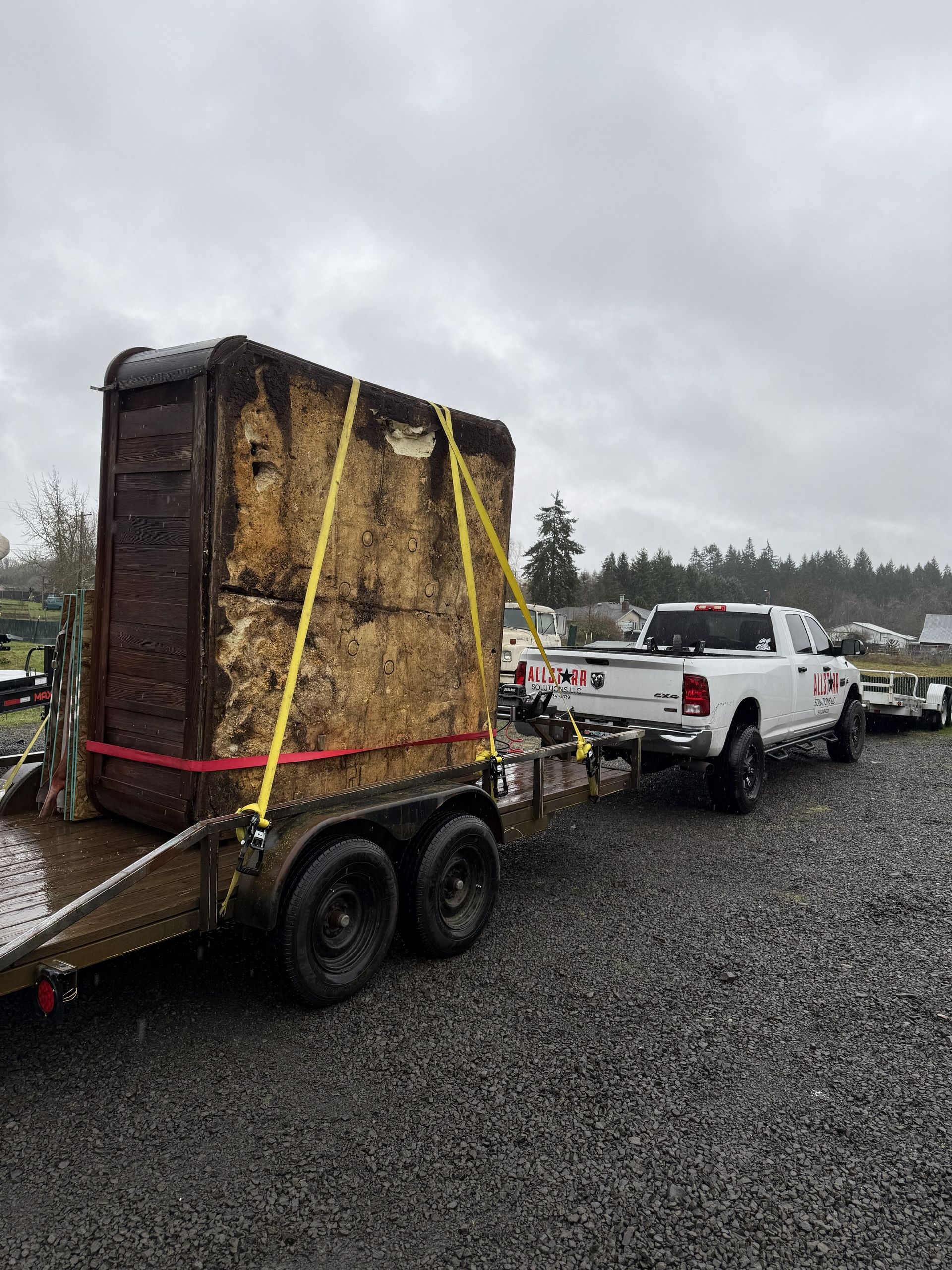 Black utility trailer carrying a large gray bin parked on a gravel lot beside a pickup truck