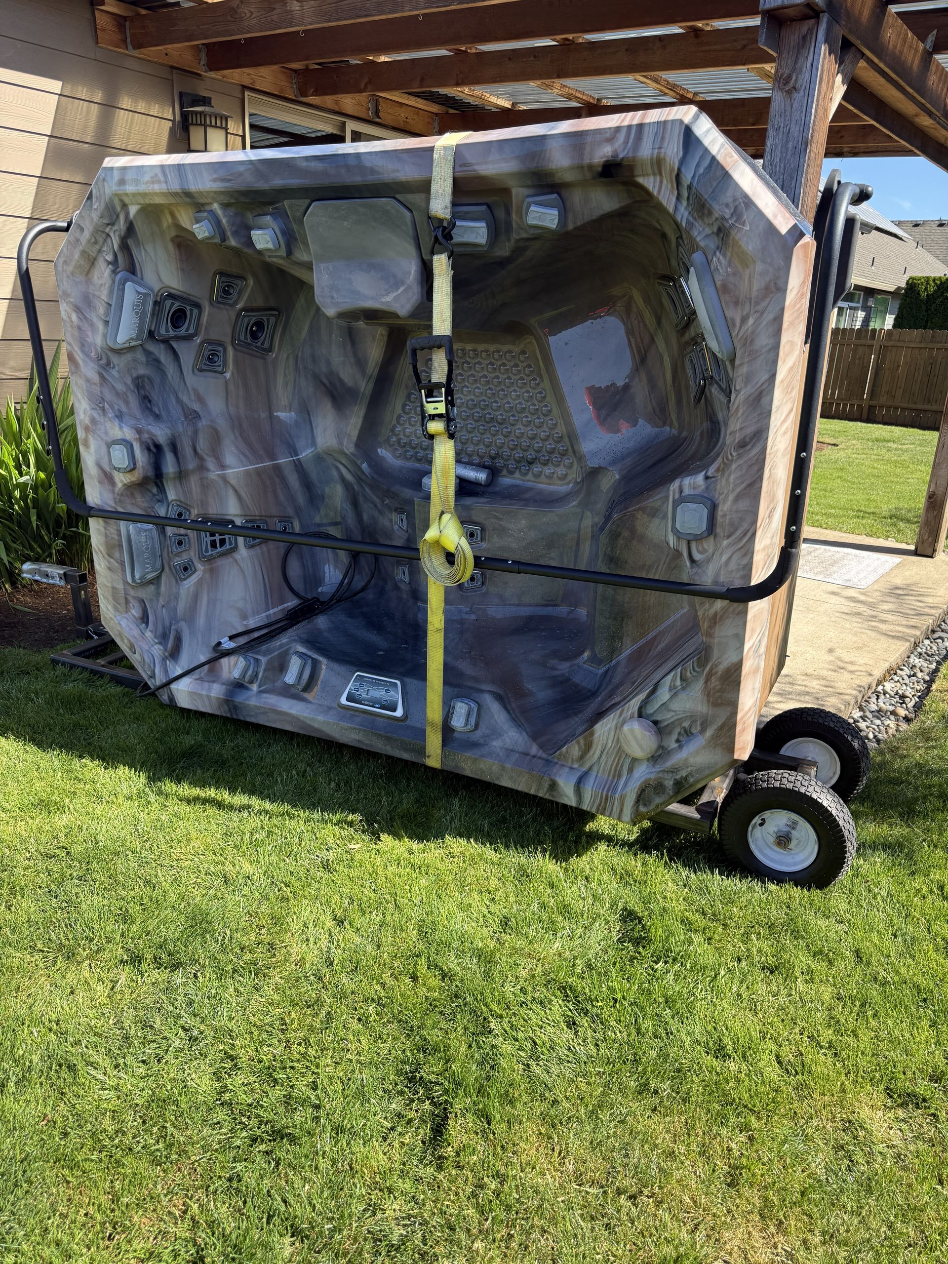 Wrapped hot tub on a wheeled dolly under a carport, secured with straps on a grassy lawn.